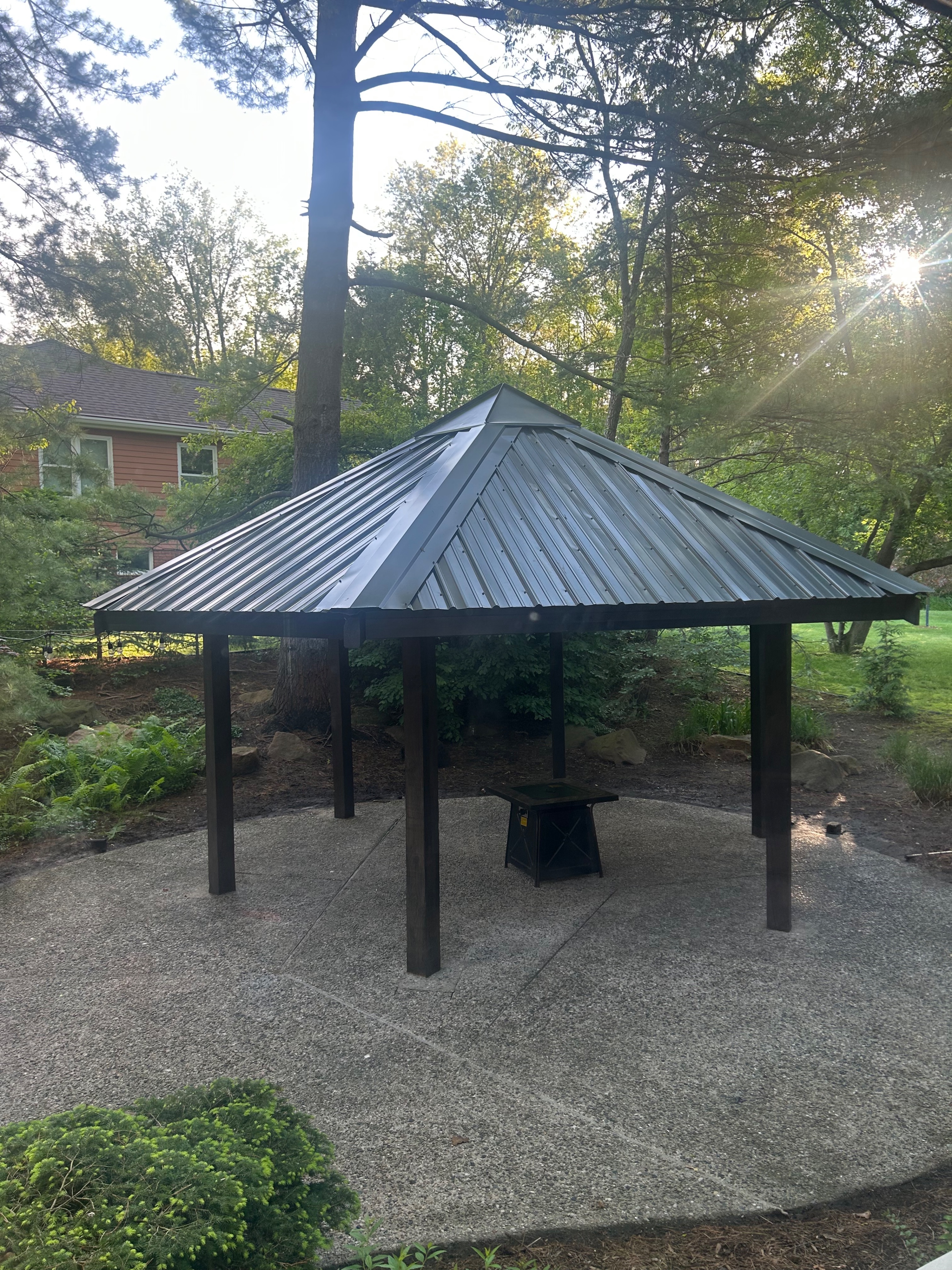 A metal gazebo with a sloped, ribbed roof and a concrete base surrounded by trees.